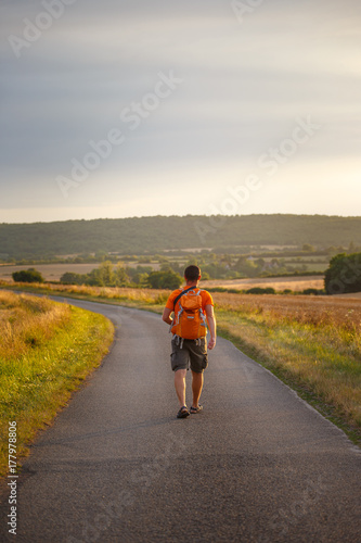 a young tourist guy walks along an asphalt road along a field in the countryside with an orange backpack and a camera around his neck in shorts and sandals in france in a burguidi in the summer