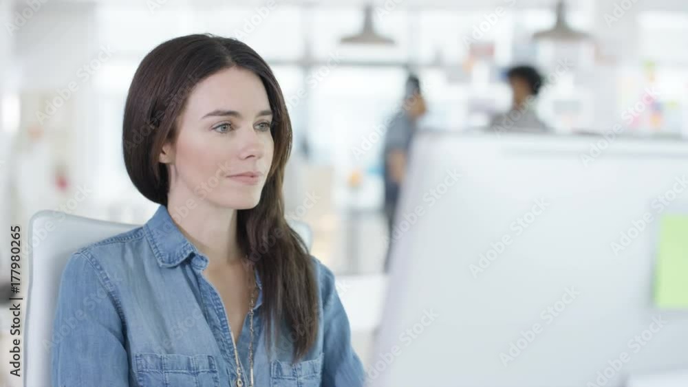 Close up young woman working on computer in creative office with colleagues talking in background. Shot on RED Epic. Close up young woman working on computer in creative office with colleagues talking in background. Shot on RED Epic.