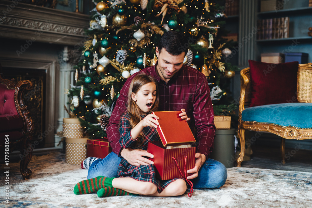 Surprised adorable small girl looks into present box, doesn`t believe her eyes, recieves pleasant gift from father, sit together on floor in cozy room near decorated New Year tree. Majestic momemt