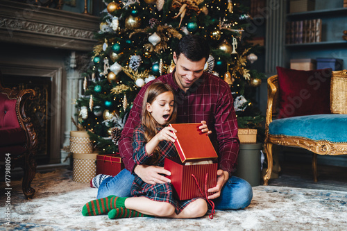 Surprised adorable small girl looks into present box, doesn`t believe her eyes, recieves pleasant gift from father, sit together on floor in cozy room near decorated New Year tree. Majestic momemt