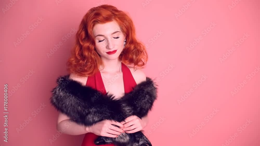 Luxurious red-haired retro woman with expensive jewelry in a red dress and winter fur cape posing on a pink background. Elegant emotional girl looking smiling and looking at the camera Luxurious red-haired retro woman with expensive jewelry in a red dress and winter fur cape posing on a pink background. Elegant emotional girl looking smiling and looking at the camera