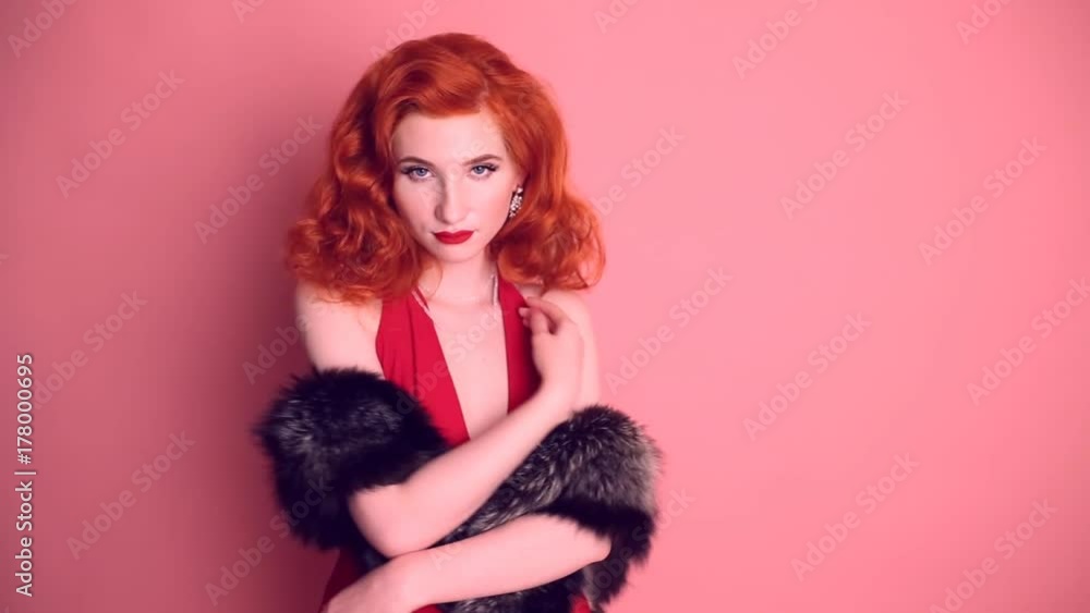 Luxurious red-haired retro woman with expensive jewelry in a red dress and fur cape posing on a pink background. Elegant emotional girl Luxurious red-haired retro woman with expensive jewelry in a red dress and fur cape posing on a pink background. Elegant emotional girl