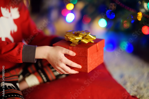 Child in pajamas holding a gift by a Christmas tree on Christmas eve