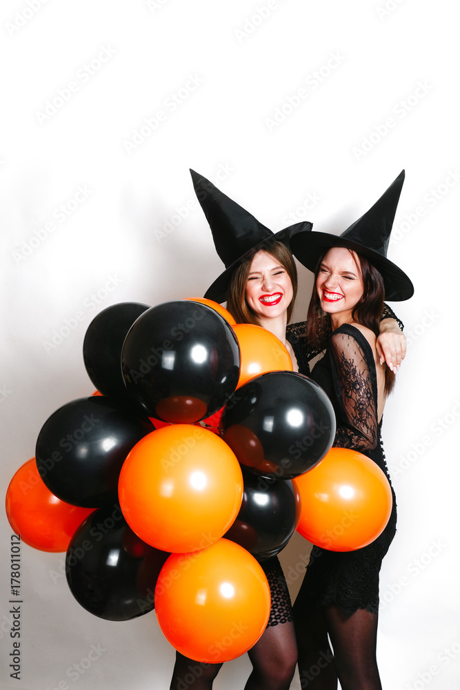 Two happy sexy women in black witch halloween costumes with orange and black balloon on party over white background