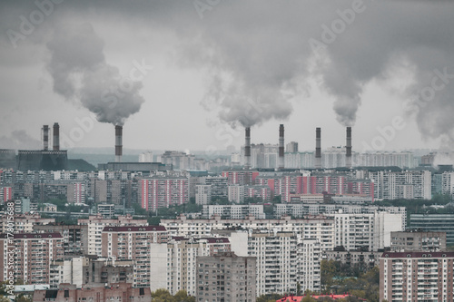 Grey and dirty residential district from high point: multiple boring dwelling houses in foreground and factory chimney-stalks in blurred background polluting atmosphere and environment with coal smoke