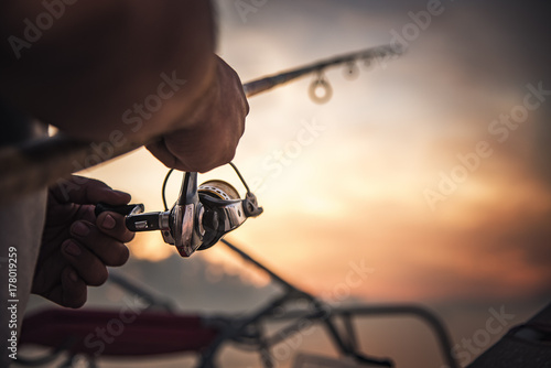 Murais de parede Fishing rod wheel closeup, man fishing with a beautiful sunrise behind him