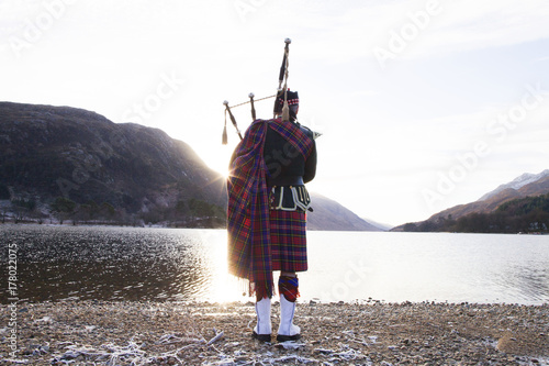 Bagpiper playing bagpipes by loch, at sunrise. Scotland. photo