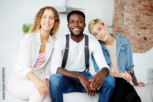 Tableau sur toile Happy young girls and African-american guy sitting on sofa