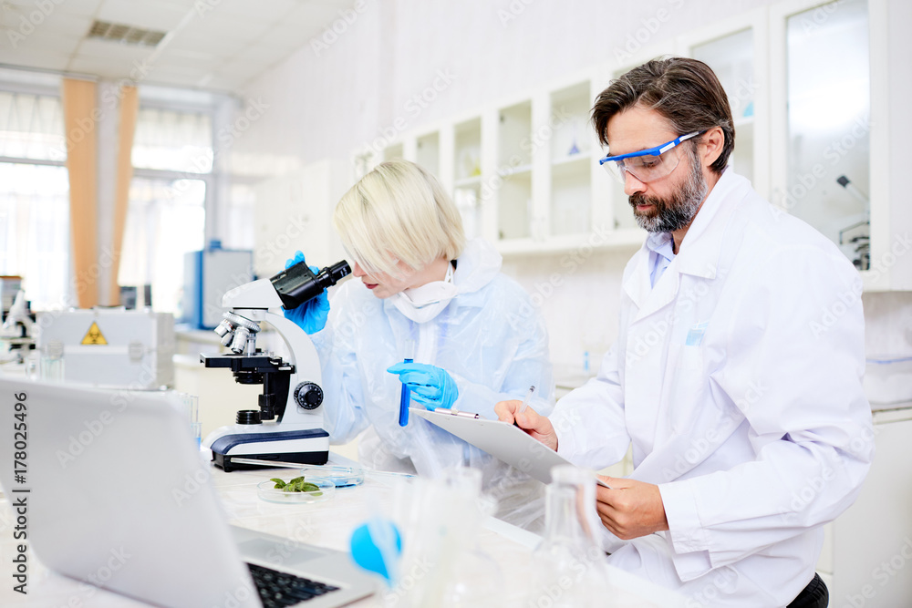 Scientist in whitecoat making notes while his colleague studying chemical substance in microscope Scientist in whitecoat making notes while his colleague studying chemical substance in microscope