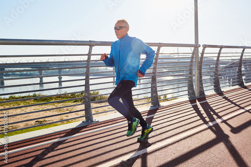 Fototapeta Naklejka Na Ścianę i Meble -  Healthy elderly man in sportswear running along athletic track while preparing for marathon, picturesque view on background, cloudless blue sky
