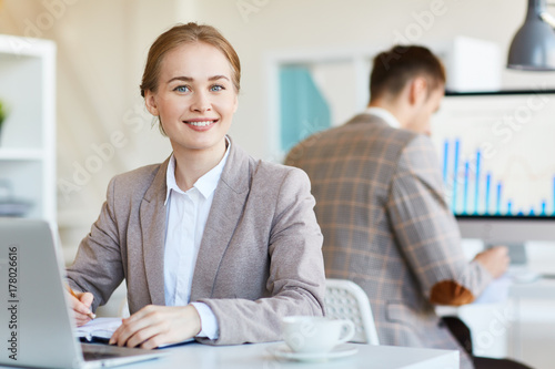 Happy businesswoman making notes in working diary with her colleague on background