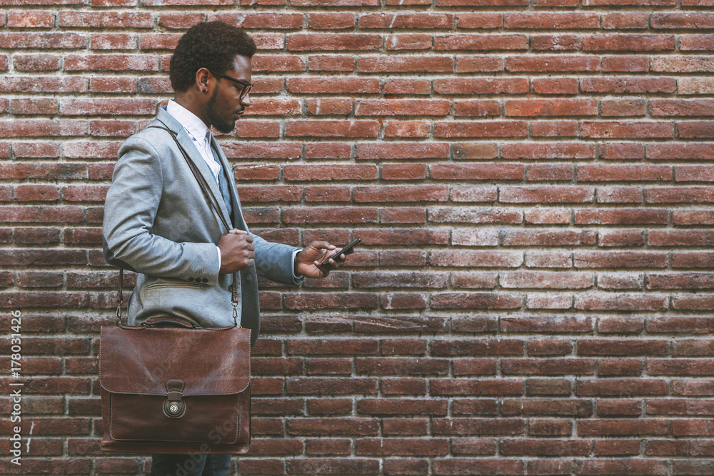 Cute black businessman sending text message with his mobile phone in front a brick wall. Cute black businessman sending text message with his mobile phone in front a brick wall.