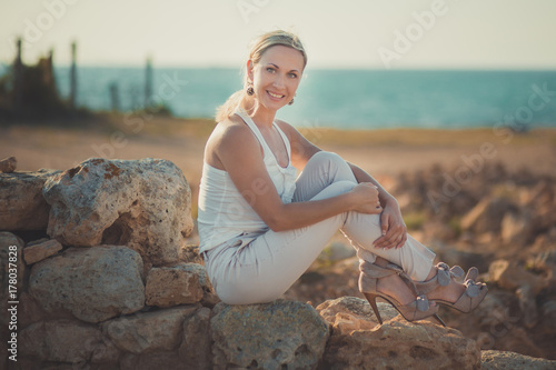 Amazing lady blond woman in light white stylish clothes sexy posing on sea side beach air.Sparkler girl looking to camera on ancient city stones blue ocean on background. photo