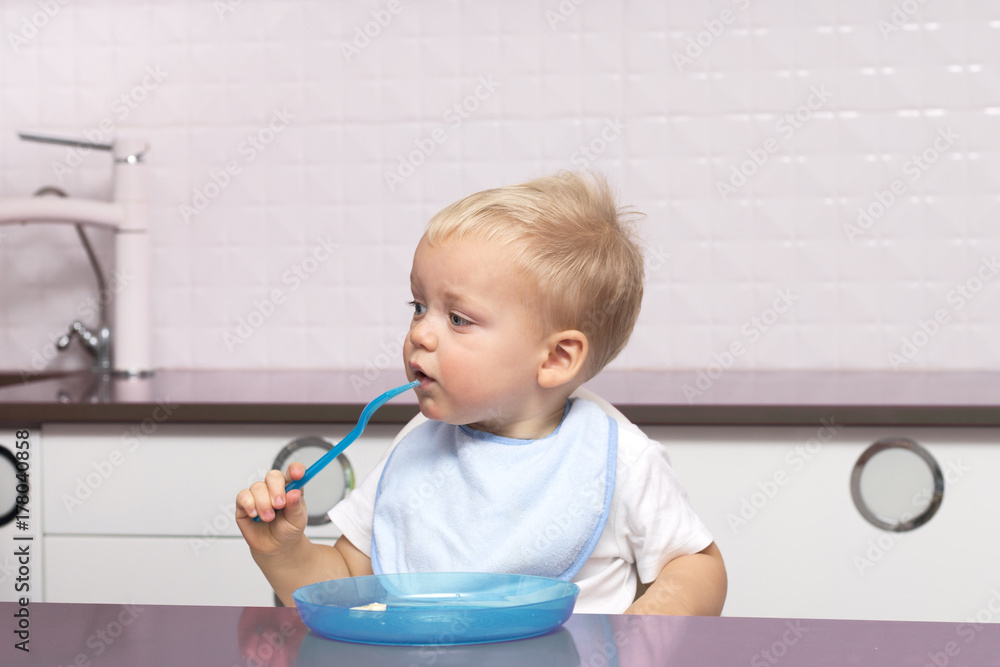 Cute toddler in a blue bib eating banana in the modern kitchen Cute toddler in a blue bib eating banana in the modern kitchen