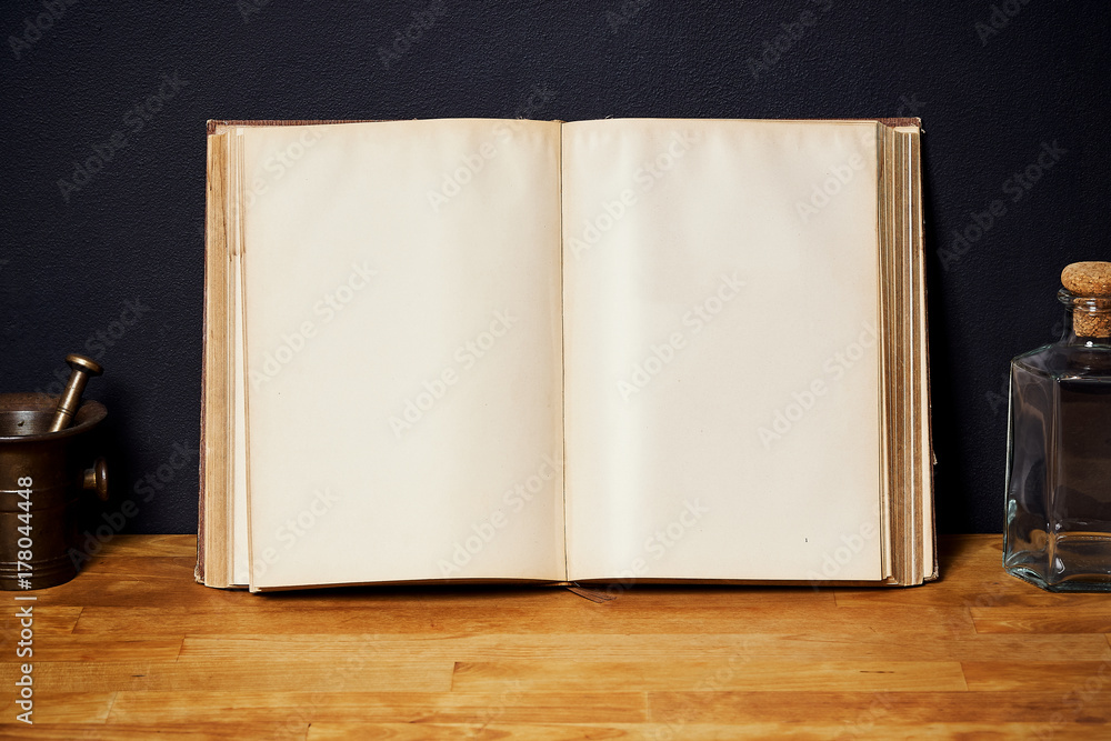 Open book on a bright wooden shelf on a black wall with a bottle and a mortar Open book on a bright wooden shelf on a black wall with a bottle and a mortar