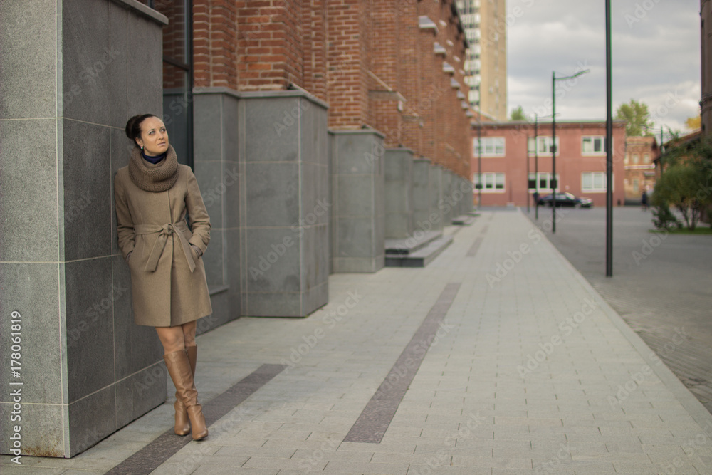 Young woman in a beige coat stands near a brick wall and laughs, does not look at the camera, red wall Young woman in a beige coat stands near a brick wall and laughs, does not look at the camera, red wall