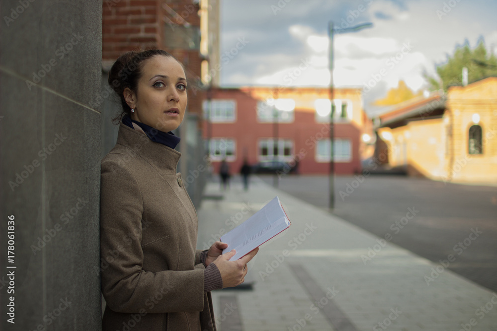 A young woman stands near a red wall, leans against a wall and reads a book, looks at the camera