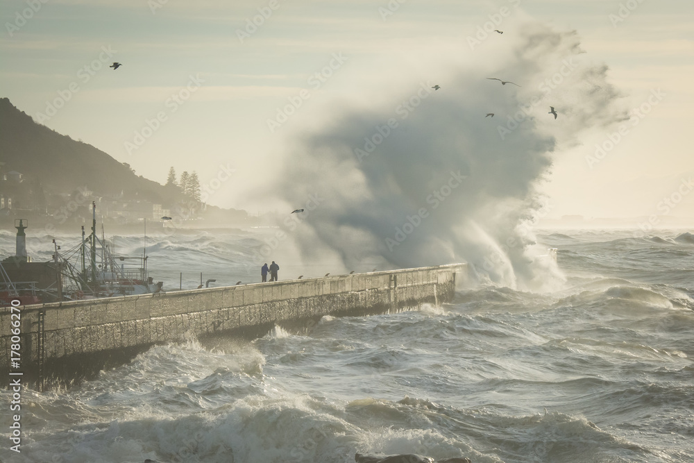 People risk their lives at Kalk Bay as a huge winter storm ocean wave crashes over the harbor wall.
