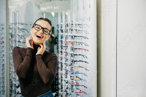 Optics. Close-up of a young beautiful girl wearing glasses, smiling near the stand. Оptical store.