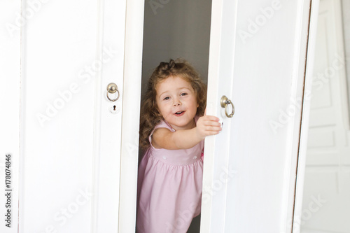 Adorable little girl hiding and looking out of wooden wardrobe. 