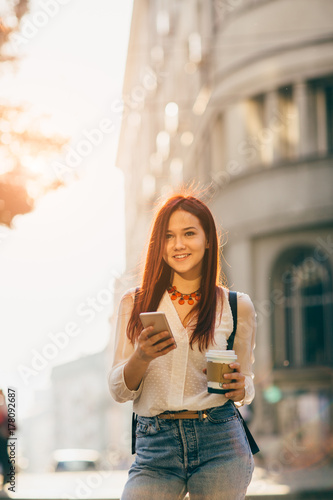 Young woman with coffee to go standing at the street and using mobile phone 