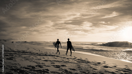 silhouette surfers walking on the beach going for surf session in scenic beautiful sunset seascape in black and white sepia, capbreton, france