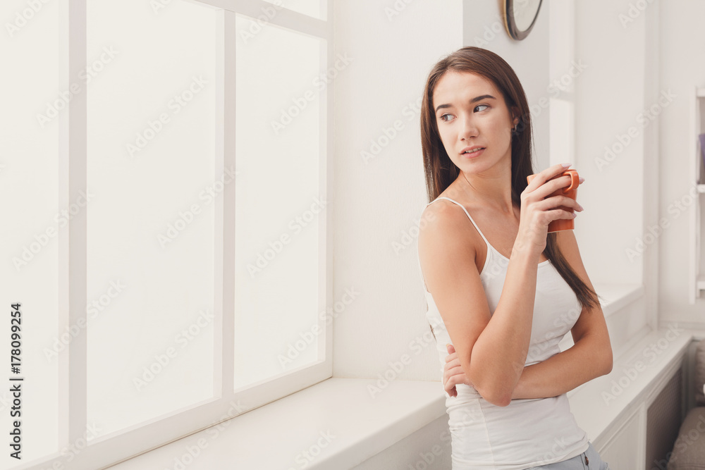 Pensive woman standing near window with coffee Pensive woman standing near window with coffee
