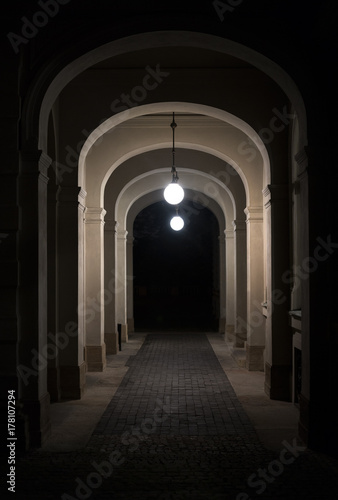 Narrow alley during night. Passage is lit by light. Lantern is on the top. Beginning and ending corridor is in the dark © M-SUR