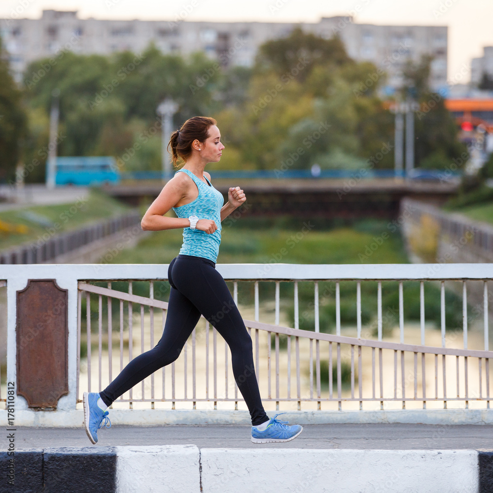 Young smiling sporty woman running on the bridge in the morning. Fitness girl jogging in city