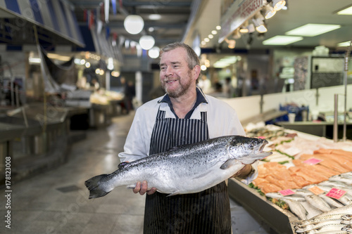 Male fishmonger wearing an apron holding large and whole salmon fish in front of display counter early in the morning on a market in England, UK.