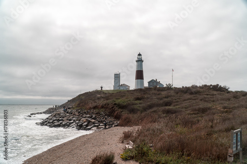 Wide establishing view of beautiful ocean coast lighthouse standing tall over rocky beach. Tourists and people fish and walk the rocky shoreline during the day