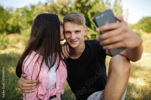 Portrait of young cool boy sitting on lawn in park and embracing girl while making selfie. Cute couple taking photos on cellphone frontal camera