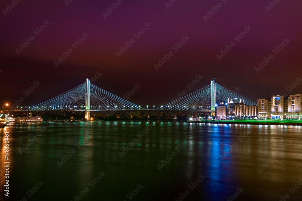 Night lights of the city are reflected in the water with a view of the bridge. Crimson night sky