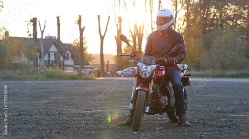 A young man in black leather jacket and white helmet sits on motorcycle turns on lights before journey at autumn sunset.