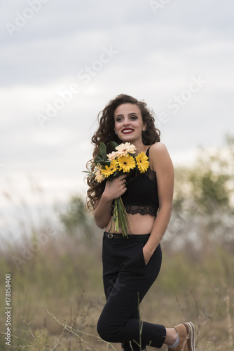 Young pretty woman with curly long hair holding a bouquet of flowers, outdoor shoot with cloudy sky