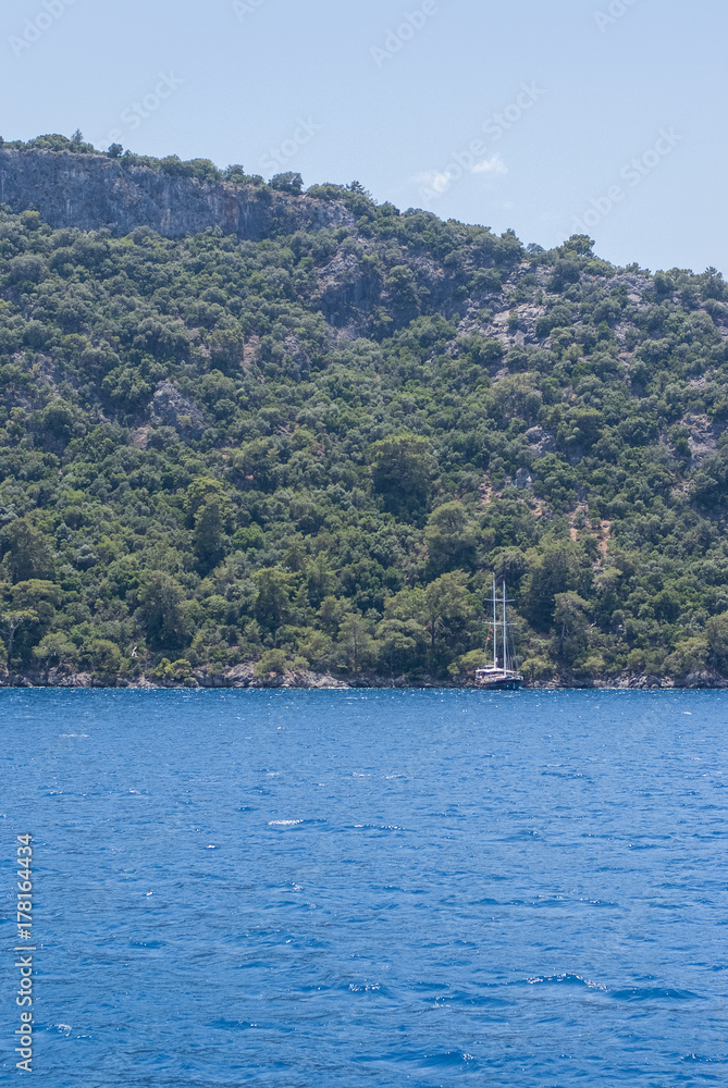 yacht with masts stands near a wooded hill in the blue sea