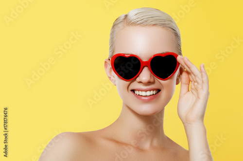 Valentine's Day concept. Fashion Model girl isolated over yellow background. Beauty stylish blonde woman posing in heart shaped sunglasses. Casual style with beauty accessories. High fashion style