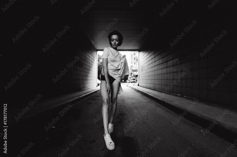 Fototapeta premium young woman with bare legs dressed in white sneakers and a long T-shirt, against the backdrop of cityscapes. street fashion, black and white style