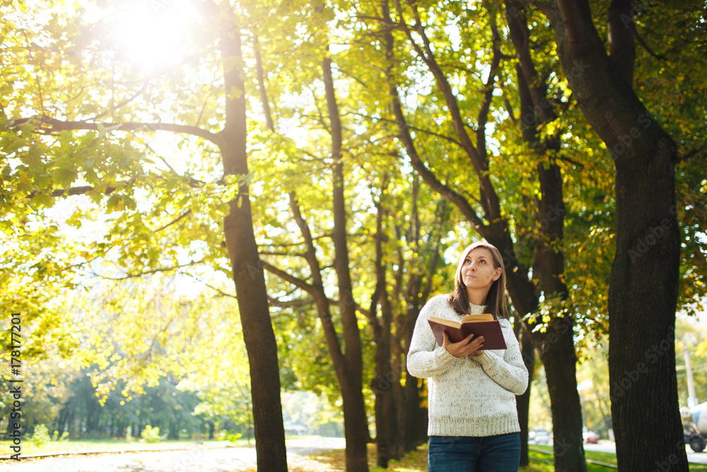 A beautiful happy smiling brown-haired woman in white sweater standing with a red book in fall city park on a warm day. Autumn golden leaves. Reading concept. A beautiful happy smiling brown-haired woman in white sweater standing with a red book in fall city park on a warm day. Autumn golden leaves. Reading concept.