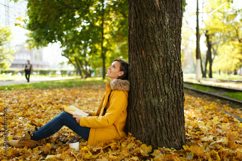 Beautiful happy brown-haired woman in yellow coat and jeans sitting under the maple tree with cup of tea or coffee and red book in fall city park. Autumn golden leaves. Reading concept.