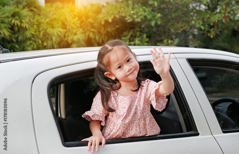 Fototapeta premium Adorable asian little girl in car smiling and looking camera sitting on a backseat of a car waving goodbye.
