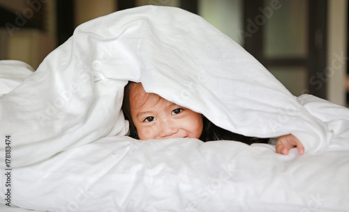 Smiling little girl lying under a white blanket on the bed.