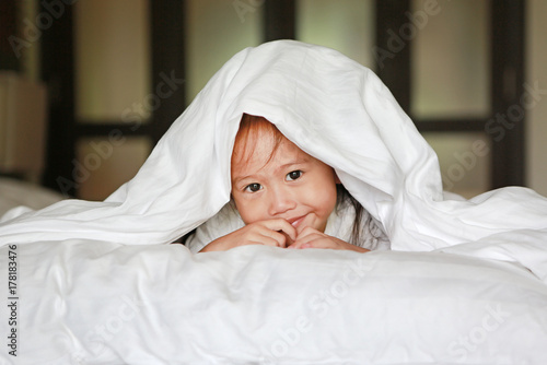 Cute little asian girl playing under a white blanket on the bed.
