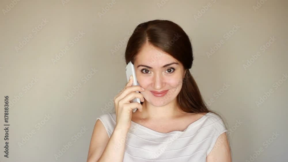 Beautiful young brunette woman talking on the phone, smiling and looking at the camera. Happy girl with a smartphone in her hand on a light background Beautiful young brunette woman talking on the phone, smiling and looking at the camera. Happy girl with a smartphone in her hand on a light background