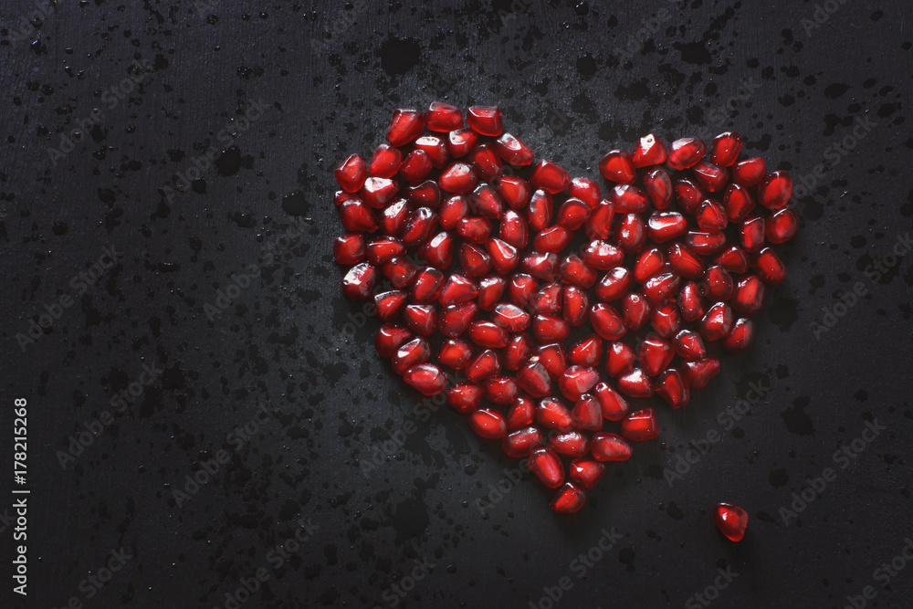 Pomegranate seeds are stacked in the form of a heart on a black background, sprinkled with water. Copy space for your text. Pomegranate seeds are stacked in the form of a heart on a black background, sprinkled with water. Copy space for your text.