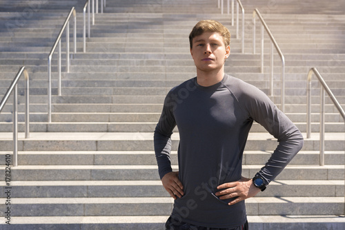 Young man in sportswear in urban place standing resting after exercises, city in sunnyday.