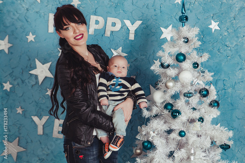 Mother holds newborn baby in sailor sweater at plastic white christmas tree against blue wall background