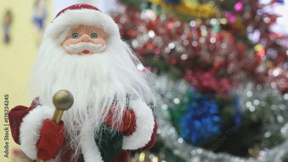 Close up of toy Santa Claus standing next to the christmas tree and slowly moves his hands in different directions in kindergarten at the New Year and Christmas holiday. Close up of toy Santa Claus standing next to the christmas tree and slowly moves his hands in different directions in kindergarten at the New Year and Christmas holiday.