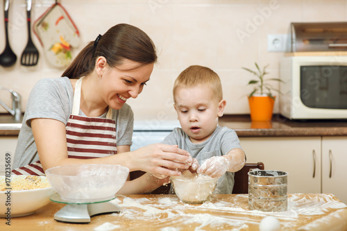 Little kid boy helps mother to cook Christmas ginger biscuit in light kitchen with tablet on the table. Happy family mom 30-35 years and child 2-3 in weekend morning at home. Relationship concept