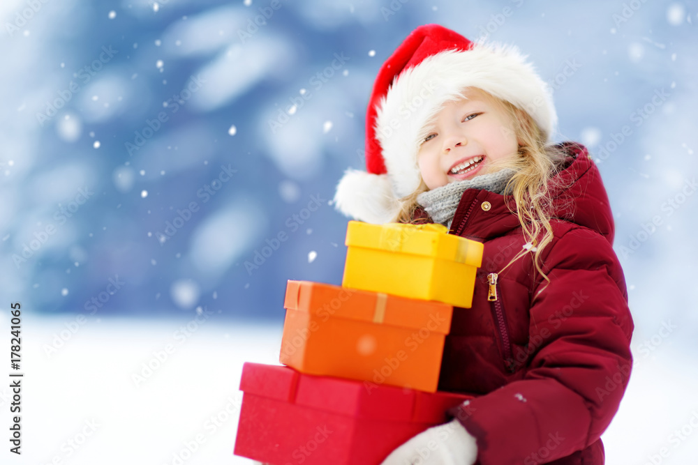 Adorable little girl wearing Santa hat holding a pile of Christmas gifts on beautiful winter day Adorable little girl wearing Santa hat holding a pile of Christmas gifts on beautiful winter day